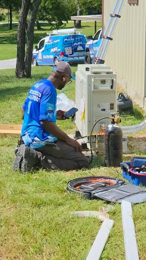 Technician working on an air conditioning unit outdoors, using a smartphone.